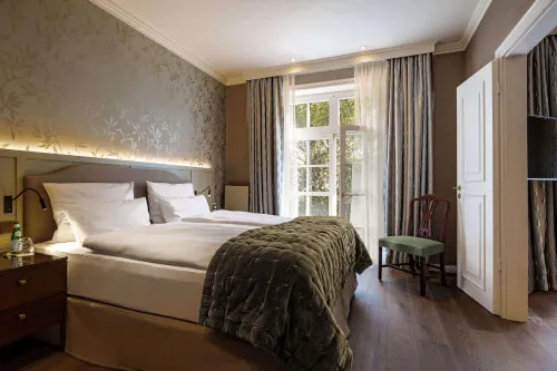 Ein komfortables Bett mit grüner Decke im Zimmer des Hotel Stadt Hamburg in Westerland, dekoriert im englischen Country-House-Stil mit Sicht auf Tür und Fenster